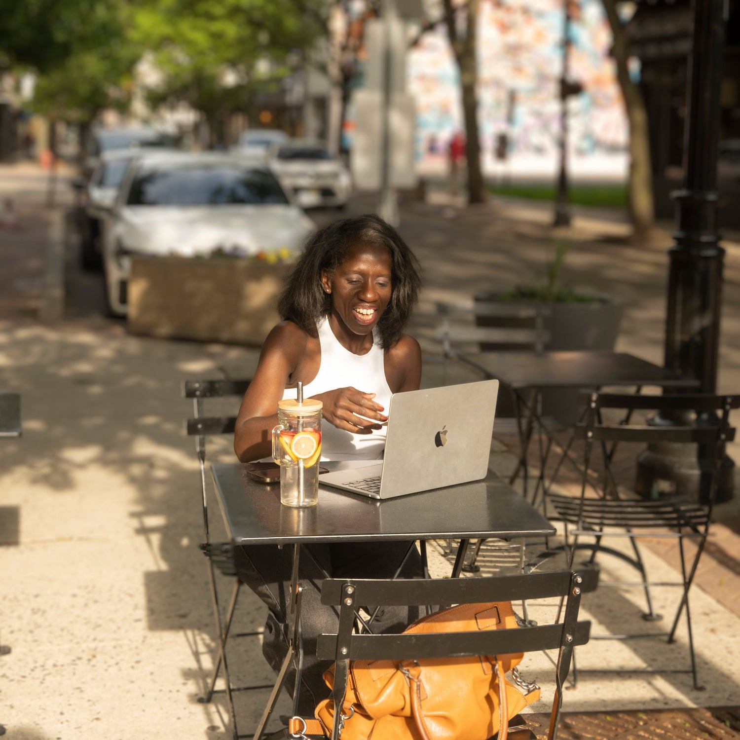 Woman using a laptop at an outdoor cafe with a yellow bag on the chair.