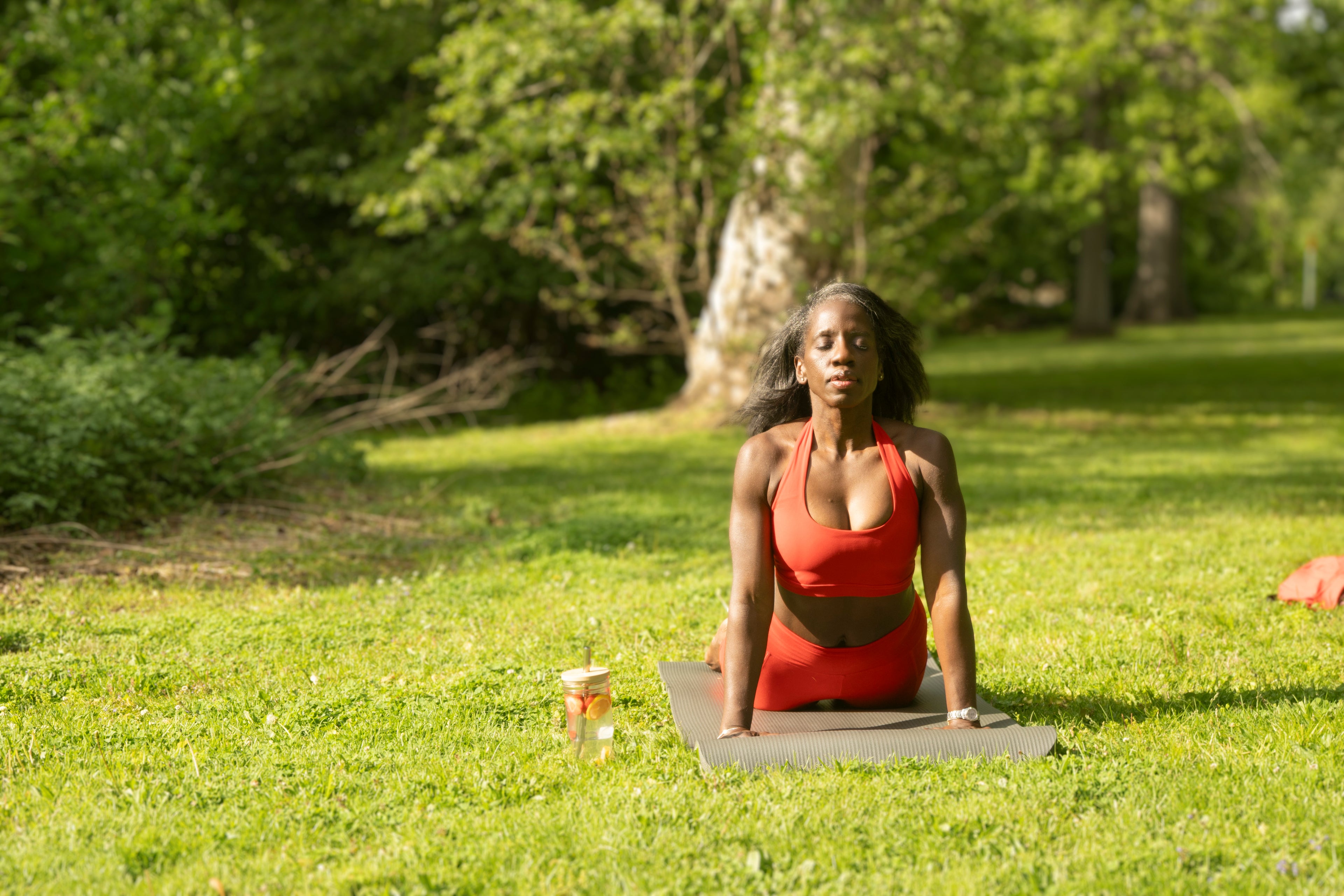 Woman in red outfit practicing yoga on a mat in a grassy outdoor area with trees in the background